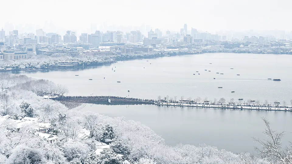 Snow-covered trees and a long causeway curve across West Lake, with Hangzhou's skyline beyond