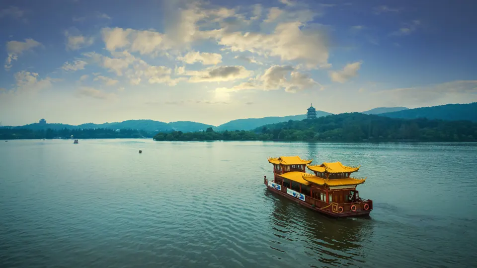 A traditional sightseeing boat crosses West Lake beneath a wide sky, with wooded hills beyond